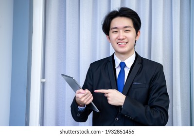 Asian Business Man Wearing Suit, Pointing To Tablet Or Smartphone. Smiling With Chat And Holding Video Call With Clients Partners.