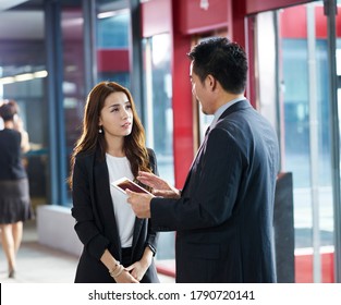 asian business man and woman standing and talking in company elevator hall using digital tablet