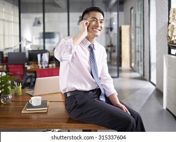 Asian Business Person Sitting On Desk Talking On Mobile Phone In Office.