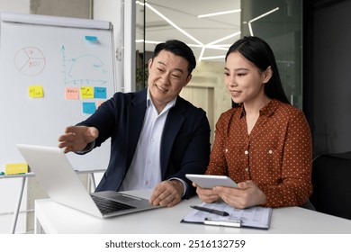 Asian business professionals engaged in collaborative work at office desk with laptop and tablet. Man explaining concepts using digital tools. Whiteboard with graphs and notes in background. - Powered by Shutterstock