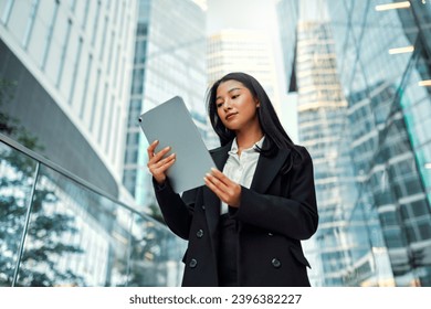 Asian business woman in a black suit holding a tablet while standing on the steps against the backdrop of business centers.