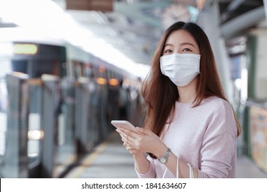 Asian business woman in casual dress code wearing face mask using mobile phone. She is waiting for the train to go to work on the platform station. New normal lifestyle in city concept