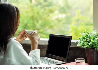 Asian business woman drinking coffee at home office enjoying the view of nature from window with laptop on desk 