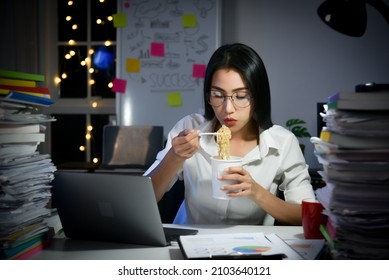 Asian Business Woman Eating Instant Cup Noodle While Working On Laptop In Office At Late Night.