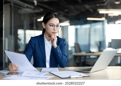 Asian business woman in office reviewing documents with laptop, showing focus and professionalism. Business attire, glasses, papers, thoughtful expression reflect corporate setting. - Powered by Shutterstock