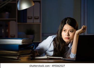 Asian Business Woman Sitting At Desk Working And Thinking Overtime Late Night. Indoors Office Background