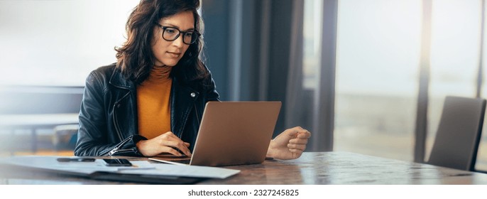 Asian business woman sitting at a table in an office, typing on her laptop with focus. Young female professional showing a dedication and commitment to her project at work. - Shutterstock ID 2327245825
