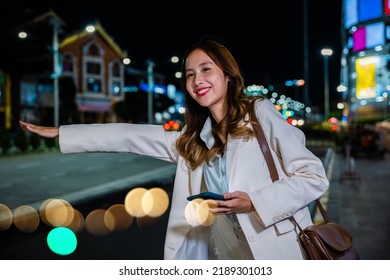 Asian Business Woman Walking To Hail Waving Hand Taxi On Road In City Street At Night, Beautiful Woman Smiling Using Smartphone Application Hailing With Hand Up Calling Cab Outdoor After Late Work