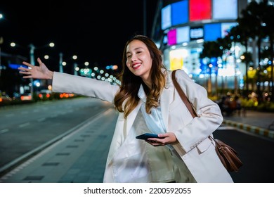 Asian Business Woman Walking To Hail Waving Hand Taxi On Road In City Street At Night, Beautiful Woman Smiling Using Smartphone Application Hailing With Hand Up Calling Cab Outdoor After Late Work