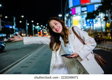 Asian Business Woman Walking To Hail Waving Hand Taxi On Road In City Street At Night, Beautiful Woman Smiling Using Smartphone Application Hailing With Hand Up Calling Cab Outdoor After Late Work