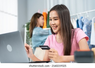 Asian Business Women Enjoy Shopping Online In Web Store In Warehouse. Young Attractive Business People Work Sit On Table, Hold Credit Card To Make E Bank Online Payment After Use Labtop Purchase Goods