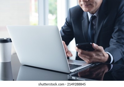 Asian Businessman In Black Suit Busy Working On Laptop Computer With Mobile Phone And Digital Tablet On Table At Office, Close Up. Man Manager Making Video Conference Using Zoom Online Meeting App