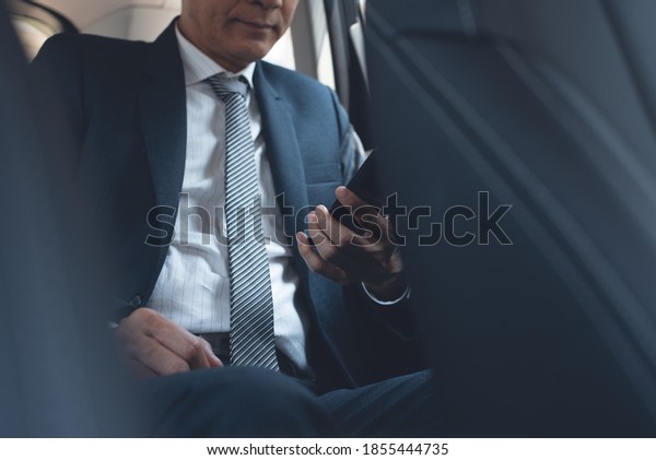 Asian businessman in black suit, executive manager\
using mobile smart phone, reading message or news application\
inside a car on backseat. Corporate man, lawyer going to workplace\
by taxi, close up