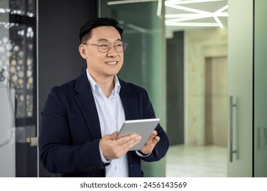 An Asian businessman in a blue blazer smiles as he uses a tablet in a well-lit, contemporary office setting, portraying professionalism and confidence. - Powered by Shutterstock