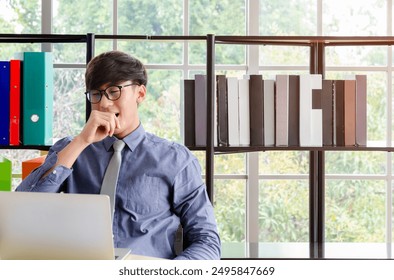 Asian businessman in blue shirt and tie sitting at desk in modern office with large windows and colorful binders and books. Young man yawning while hard working with laptop - Powered by Shutterstock