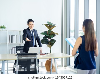 Asian Businessman, Boss In Suit Making Hand Gesture To Invite Young Woman Holding Resume In The Job Interview To Sit Down On The Chair Near Huge Glass Window In Manager Room In Modern Office Interior.