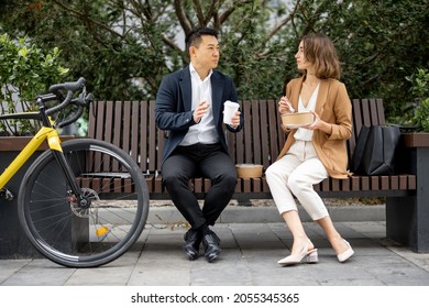 Asian Businessman And Caucasian Businesswoman Eating Food And Drinking Coffee While Having Lunch At Work. Concept Of Rest And Break On Job. Business People Sitting On Bench On City Street