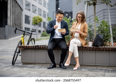 Asian Businessman And Caucasian Businesswoman Eating Food And Talking While Having Lunch At Work. Concept Of Rest And Break On Job. Smiling Business People Sitting On Bench In City