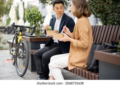 Asian Businessman And Caucasian Businesswoman Eating Food And Drinking Coffee While Having Lunch At Work. Concept Of Rest And Break On Job. Business People Sitting On Bench On City Street