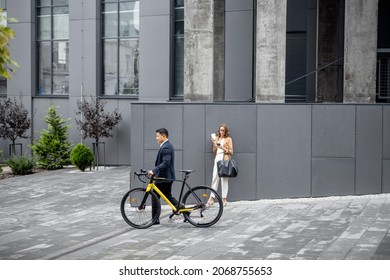 Asian Businessman And Caucasian Businesswoman On City Street. Asian Guy Passing By With A Bicycle, Woman Using Phone. Business People During A Break Outdoors. Wide View On The Office Building