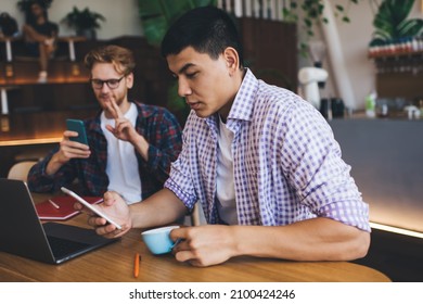 Asian Businessman Drinking Tea And Using Smartphone While His Caucasian Male Colleague Having Video Call On Mobile Phone. Business Cooperation And Teamwork. Young Men Working At Table In Office