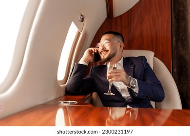 Asian businessman flies in private luxury jet and talks on the phone and drinks champagne, Korean entrepreneur in suit and glasses relaxes and communicates via mobile communication in airplane - Powered by Shutterstock