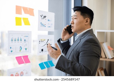 Asian businessman in formal suit stands by window analyzing charts and graphs while speaking on smartphone. Focused on business growth and data analysis. - Powered by Shutterstock