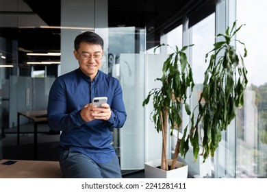 Asian businessman inside the office in a casual shirt and glasses is using the phone, the boss is holding a smartphone in his hands reading news and typing a message smiling. - Powered by Shutterstock