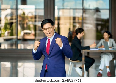 Asian businessman manager in navy blue formal suit raises fist in joy with team behind him outside in an off-site working concept. - Powered by Shutterstock