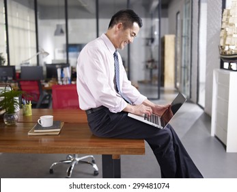 An Asian Businessman Sitting On Desk Using Laptop Computer In Office, Happy And Smiling, Side View.