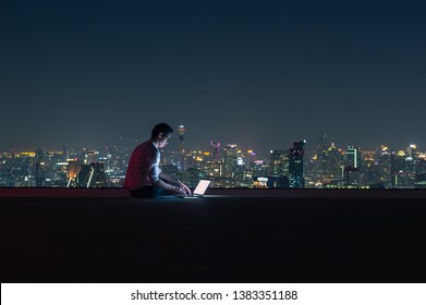 Asian Businessman Sitting At The Top Of The Building. Use The Laptop Computer With The Scenery Of The Night View Of The City, Business Success And Technology Concept.