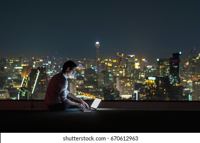 Asian Businessman Sitting And Using The Laptop Over The Cityscape Background At Night Time, Business Success And Technology Concept