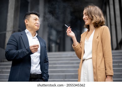 Asian businessman and smiling caucasian businesswoman smoking cigarettes during break at work on city street. Concept of modern successful business people. Idea of rest and break on job - Powered by Shutterstock