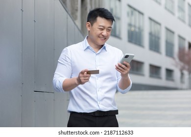 Asian businessman smiling and happy doing online shopping outside office building, man using mobile phone, holding bank credit card - Powered by Shutterstock