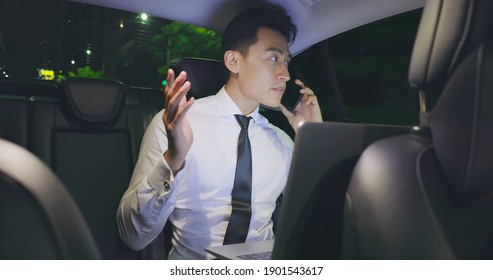 Asian Businessman Speaks On Smart Phone And Uses Laptop Computer To Work In Car While Commuting At Night