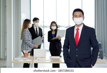 An Asian Businessman In A Suit Stands In His Pocket With A Confident Expression. The Team Is Preparing For The Meeting In The Background. Everyone Wears A Mask. Concept Of Leadership.