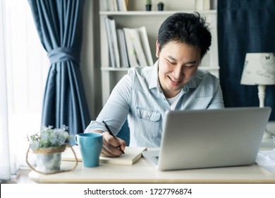 Asian Businessman Talking To Colleague Team In Video Call Conference Writing Note On Book With Smile Face. Man Using Computer Laptop For Digital Online Meeting Network. Smart Working From Home Concept