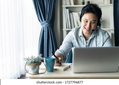 Asian businessman talking to colleague team in video call conference writing note on book with smile face. Man using computer laptop and headphone for online meeting. Smart working from home concept.