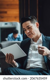 Asian Businessman Talking On Video Call Meeting In A Cafe. 
Smiling Business Man In Blue Suit Attending On Online Web Conference On A Digital Tablet While Drinking A Cup Of Coffee.