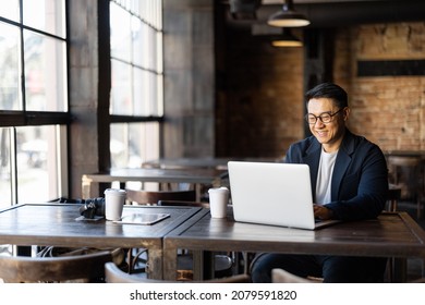 Asian businessman typing on laptop during work in cafe. Concept of remote and freelance work. Smiling adult successful man wearing suit and glasses sitting at wooden desk. Sunny day - Shutterstock ID 2079591820