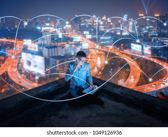 Asian Businessman Using Phone And Laptop On Rooftop With Social Connection Lines In Smart City At Night