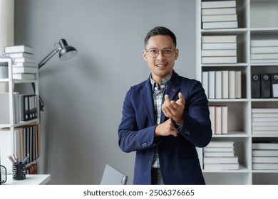 Asian businessman wearing a blue blazer over a plaid shirt is gesturing with his hand while smiling in an office - Powered by Shutterstock