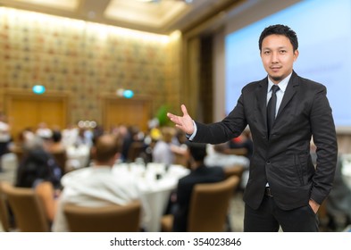 Asian Businessman with welcoming gesture on Abstract blurred photo of conference hall or seminar room with attendee background