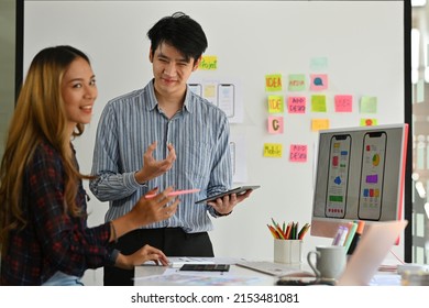 Asian Businessman And Woman Talking About New Media Application Design As Shown On The Computer Desktop Screen With Modern Office Decorated Interior. Technology And Design Concepts.