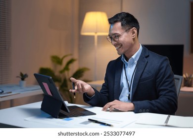 Asian businessman is working late in the office and is smiling while having a video call on his tablet - Powered by Shutterstock