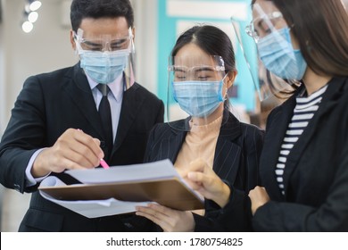 Asian businesspeople wear masks and face shield protect against airborne disease and salivary infections, during outbreak of Covid 19 virus (Coronavirus) reading reports of workers in office.