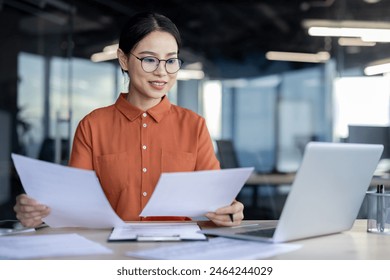 Asian businesswoman analyzing documents at desk in a modern office while working on a laptop. Professional, focused, and productive work environment. - Powered by Shutterstock