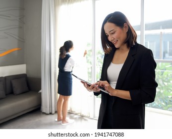 Asian Businesswoman In Black Suit Standing With Tablet Computer In Her Hand. The Hotel Manager Verify The Tidiness Of The Room For Hotel Guests While The Maid Cleaning The Curtain In The Background.