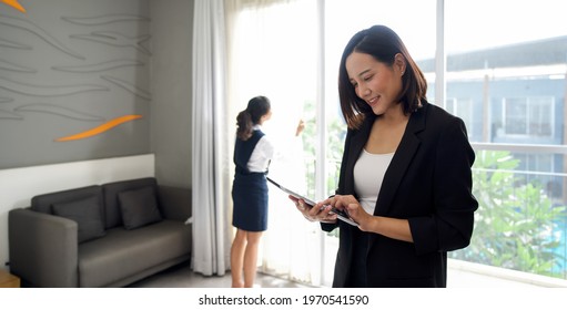 Asian Businesswoman In Black Suit Standing With Tablet Computer In Her Hand. The Hotel Manager Verify The Tidiness Of The Room For Hotel Guests While The Maid Cleaning The Curtain In The Background.