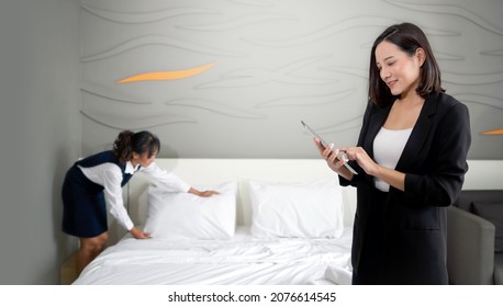Asian Businesswoman In Black Suit Standing With Tablet Computer In Her Hand. The Hotel Manager Verify The Tidiness Of The Room For Hotel Guests While The Maid Making The Bed In The Background.